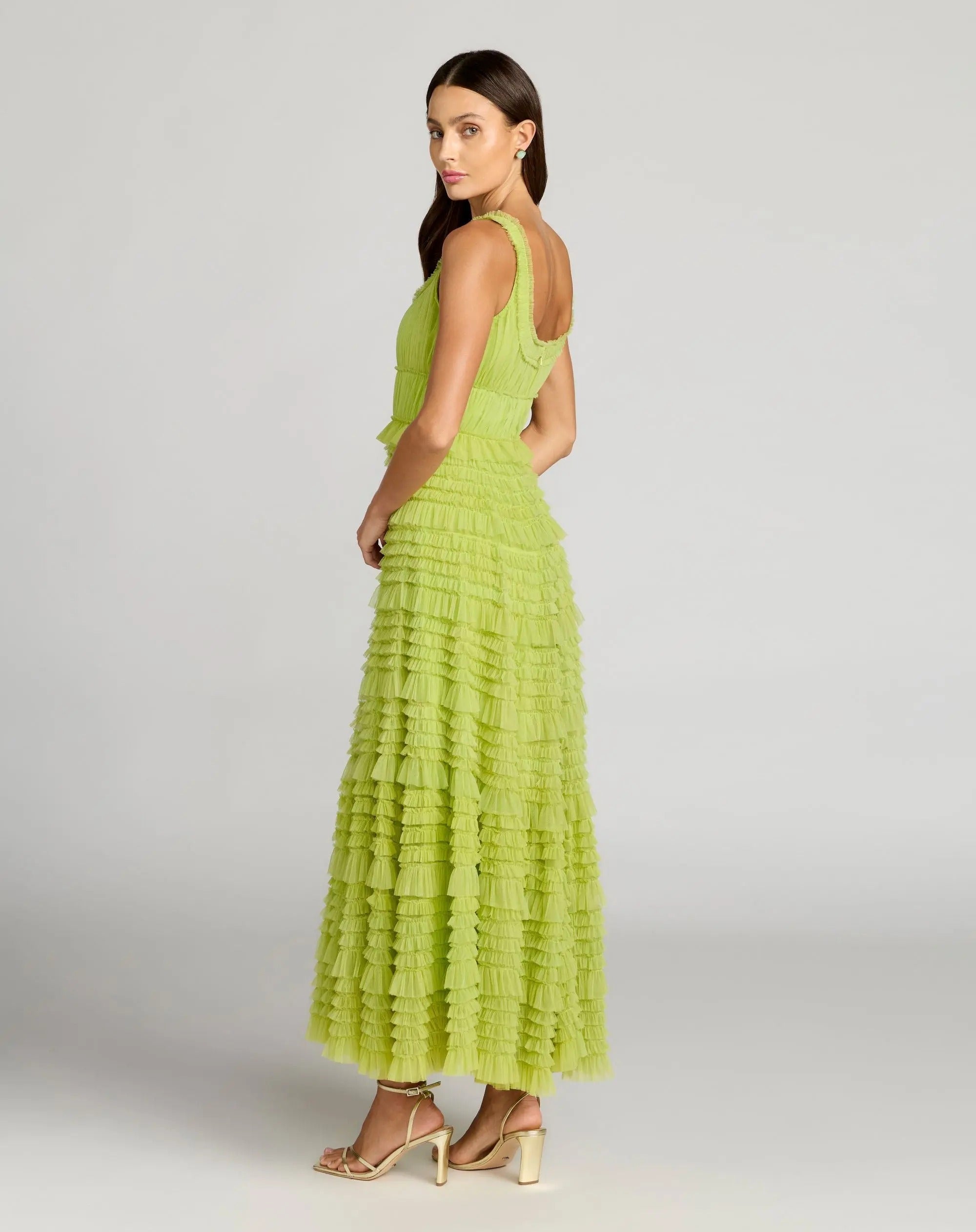 A woman stands in profile against a plain gray background, wearing the Mac Duggal 8292 tea-length party dress—a sleeveless lime green gown featuring ruffle detailing and an elegant A-line tiered skirt—paired with beige open-toe heels.